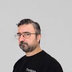 Bearded man wearing eyeglasses and a slogan sweater, looking thoughtful indoors.